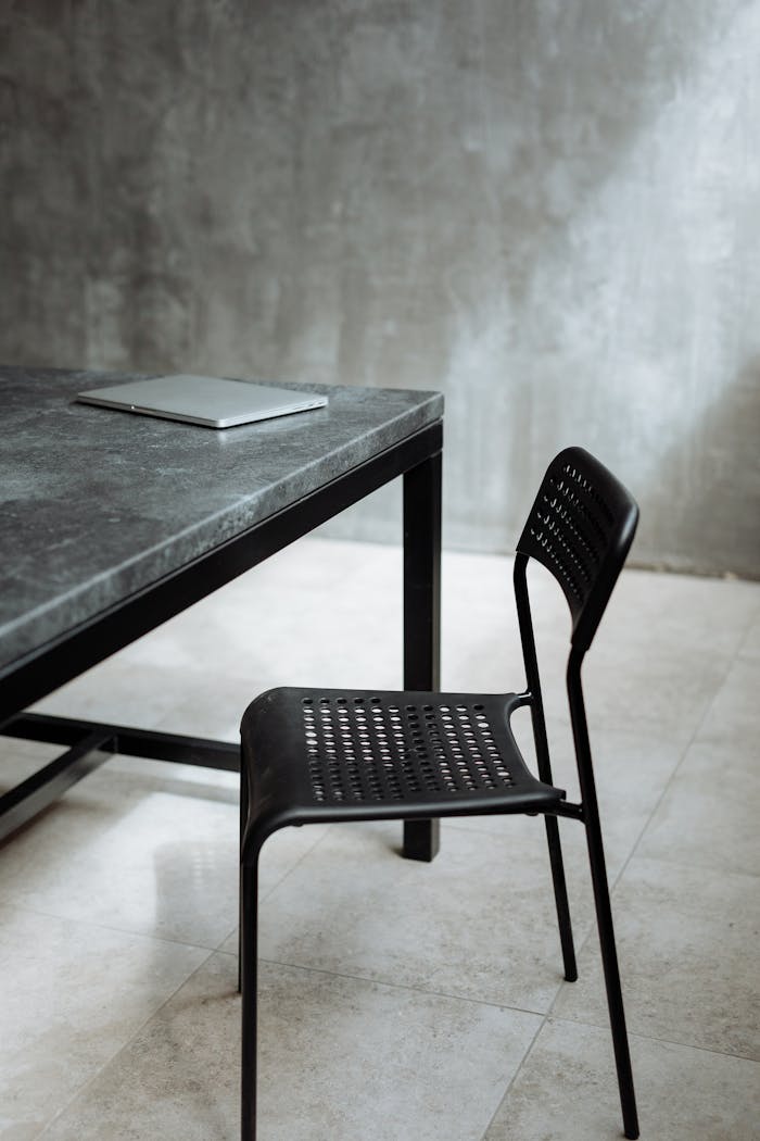 A sleek black metal chair and laptop on a marble top table against a concrete wall.