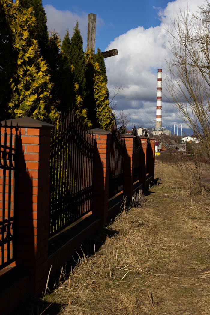 A scenic view of an industrial chimney behind a red brick fence lined with trees.